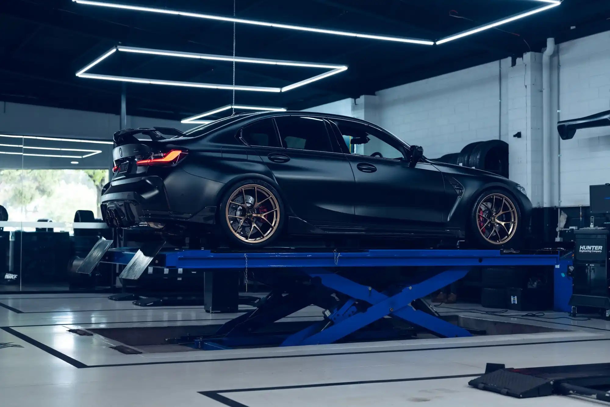 A matte black sports sedan with custom bronze wheels on a blue hydraulic lift.