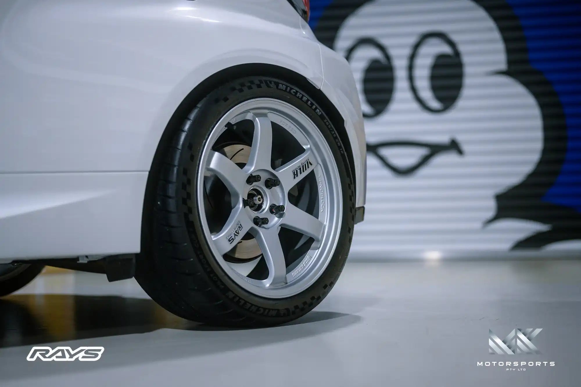 A shiny silver multi-spoke wheel with a black tire mounted on a white car.