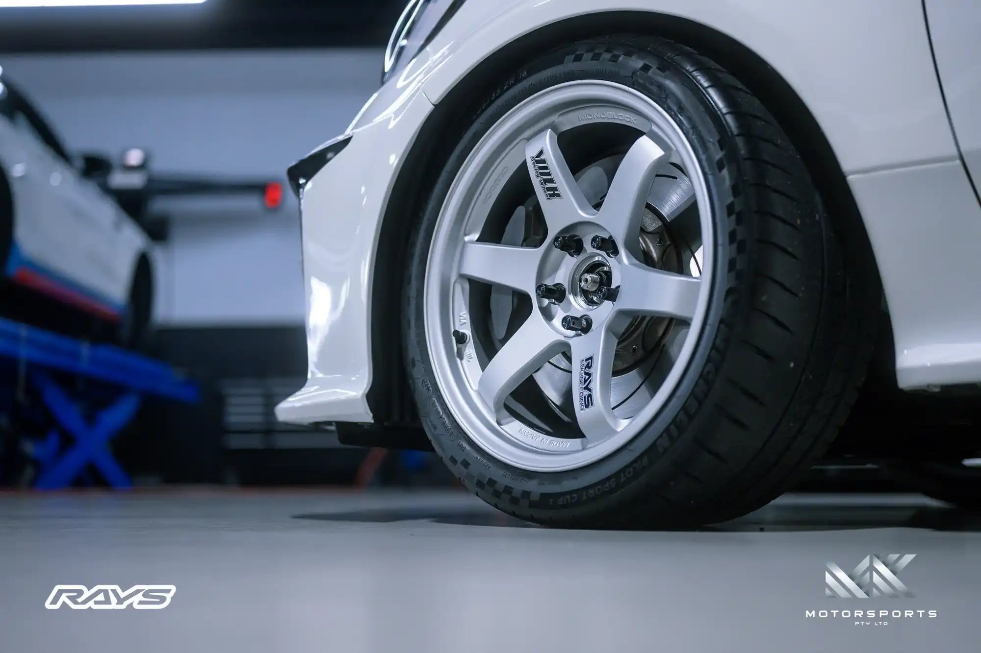 A white car with a silver, six-spoke wheel and performance tire.